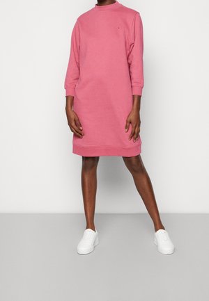 Personne portant une robe rose à manches longues, longueur genou, associée à des baskets blanches, debout devant un fond clair uni.