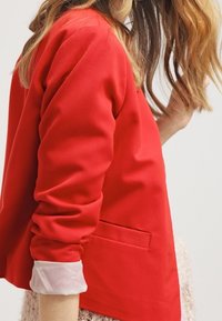 Czerwony żakiet o zdefiniowanej sylwetce, z długimi rękawami i jedną kieszenią na piersi. Posiada podwinięte białe mankiety oraz gładką tkaninę.
