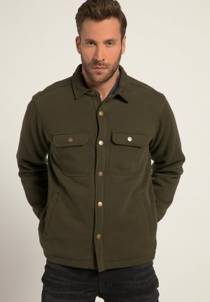 Olijfgroene jas van een gladde, gestructureerde stof, voorzien van twee borstzakken, drukknoopsluitingen en een kraag. Gedragen met donkere jeans.