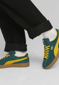 Semelle en caoutchouc pour sneakers en daim de couleur bleu canard et jaune. Comprend des rayures jaunes sur les côtés et des lacets jaunes, assortis à un pantalon noir retroussé et des chaussettes blanches.