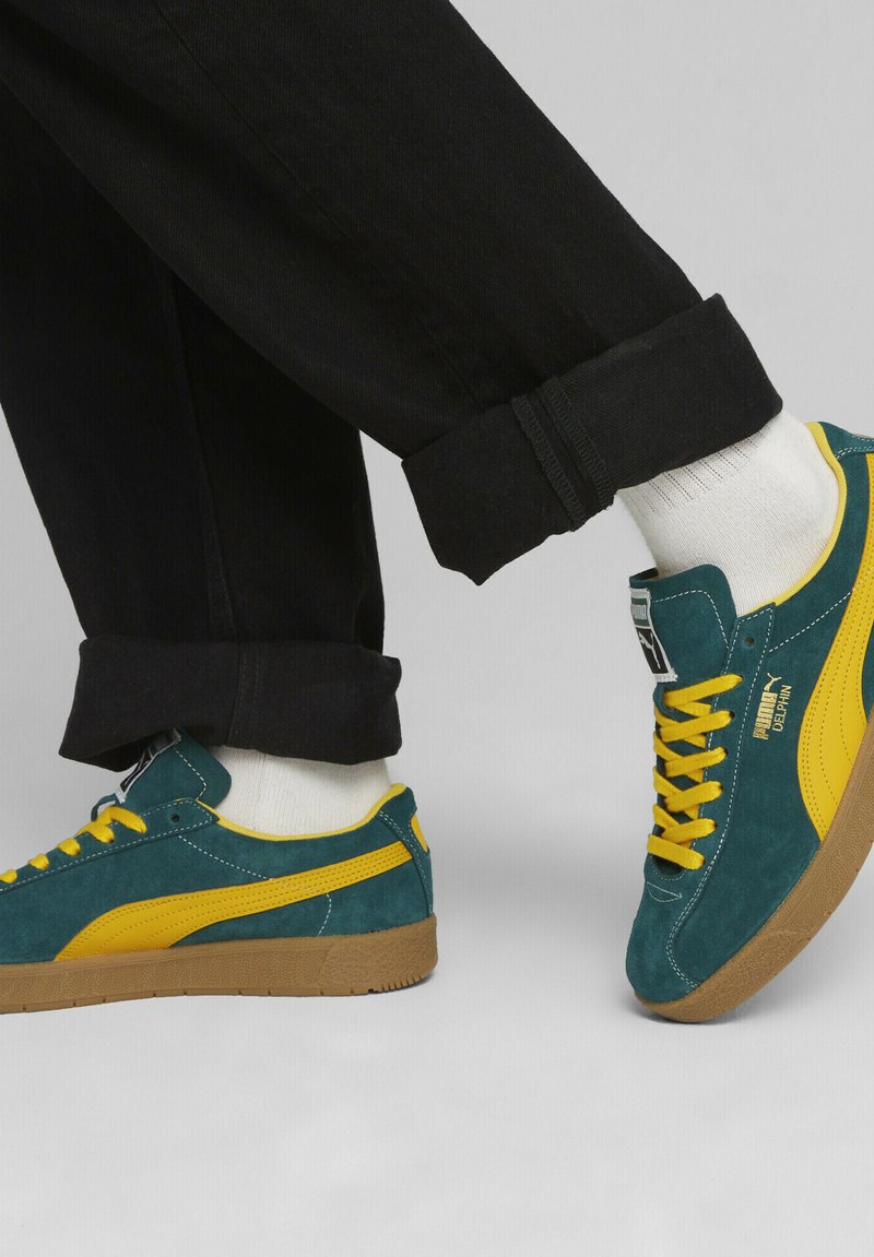 Semelle en caoutchouc pour sneakers en daim de couleur bleu canard et jaune. Comprend des rayures jaunes sur les côtés et des lacets jaunes, assortis à un pantalon noir retroussé et des chaussettes blanches.