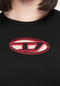 Black shirt featuring a large oval embroidery in red and black with a cut-out section at the center. Smooth texture and fitted design.