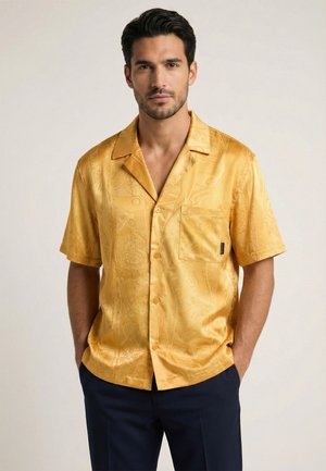 Mand med kort mørkt hår og skæg iført en sennepsgul kortærmet skjorte med mønster og mørke bukser, hænderne i lommerne.