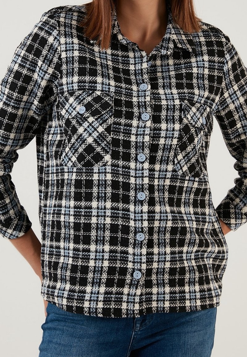 Camicia a quadri in bianco e nero, realizzata in tessuto texturizzato, con bottoni blu, due tasche sul petto e un'apertura frontale con bottoni e maniche lunghe.