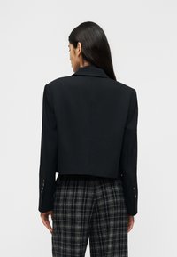 Γυναίκα με μακριά σκούρα μαλλιά που φορά ένα κοντό μαύρο σακάκι και ασπρόμαυρο παντελόνι με καρό σχέδιο, στέκεται μπροστά σε μονόχρωμο φωτεινό φόντο.