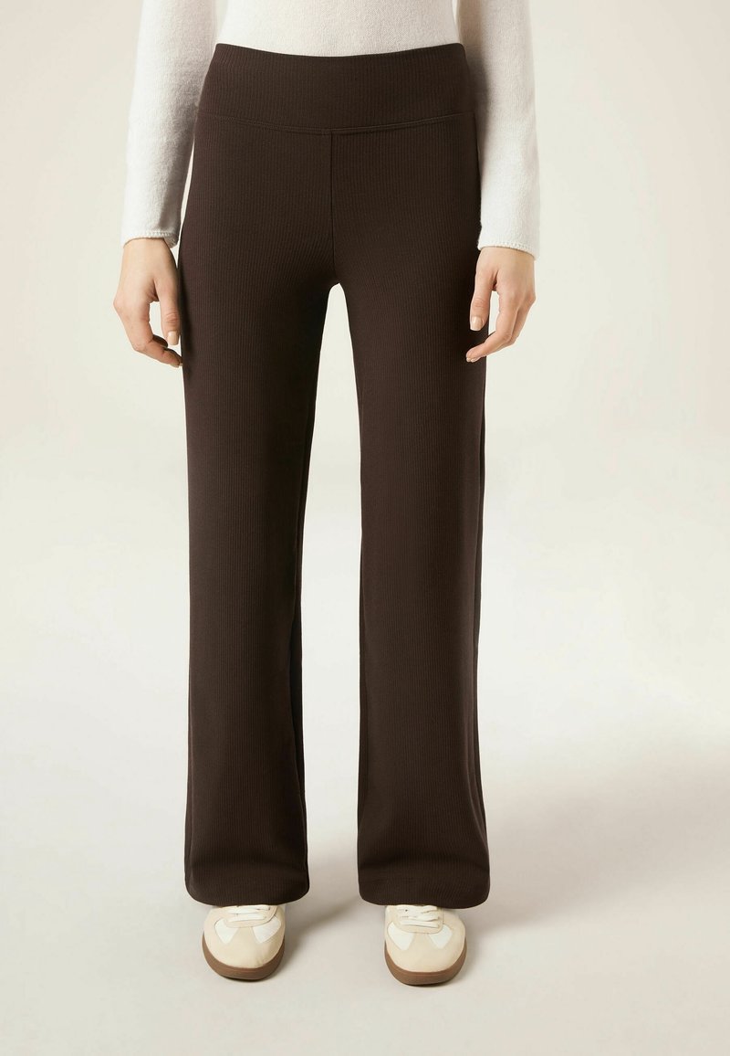 Personne portant un pantalon large côtelé marron foncé, des baskets blanches et un haut blanc à manches longues sur un fond uni.