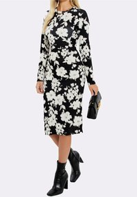 Svart långärmad klänning med vitt blommönster, kroppsnära design, knä-lång, kombinerad med svarta ankelboots och en svart handväska.