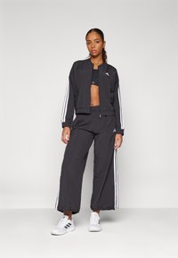 Giacca sportiva nera con strisce bianche sulle maniche, abbinata a pantaloni morbidi in coordinato. Entrambi hanno un tessuto liscio e un design corto.