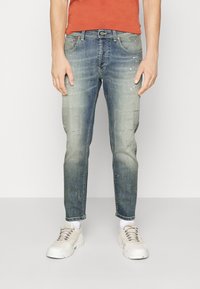 Schmal geschnittene Denim-Jeans in verblasstem Blau mit subtilen Abnutzungen, ausgefransten Akzenten und leichten Farbspritzern, kombiniert mit weißen Sneakers.