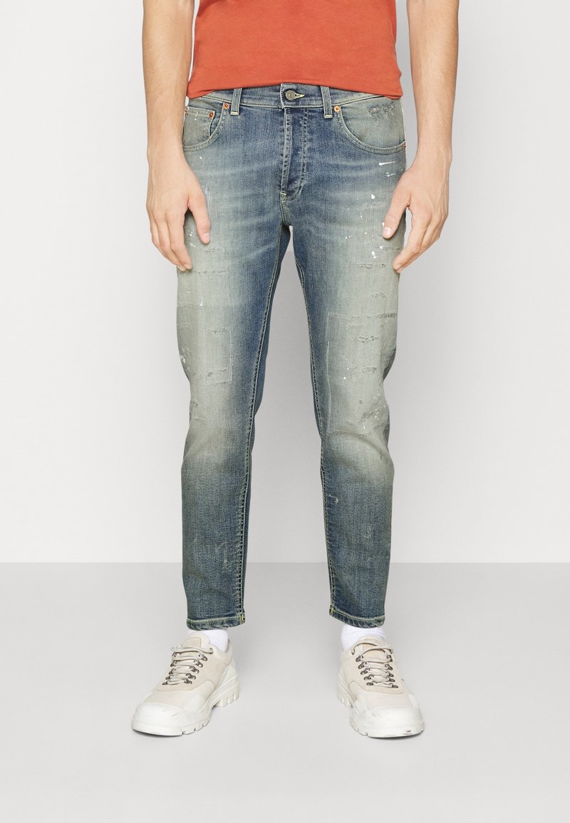 Schmal geschnittene Denim-Jeans in verblasstem Blau mit subtilen Abnutzungen, ausgefransten Akzenten und leichten Farbspritzern, kombiniert mit weißen Sneakers.