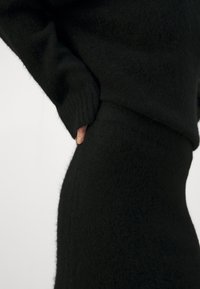 Gros plan d'une personne portant un pull noir texturé à manches longues et une jupe assortie, avec une main glissée dans la taille.