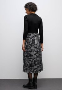 Schwarzes Oberteil mit langen Ärmeln, kombiniert mit einem knielangen grauen Rock mit einem schwarzen Zebra-Muster. Das Model trägt schwarze Knöchelstiefel.