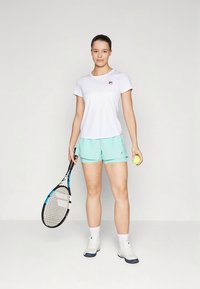 Valge lühikeste varrukatega spordisärk diskreetse logoga, kaasas mündirohelised lühikesed püksid. Modell hoiab tennisesüüdist ja palli.