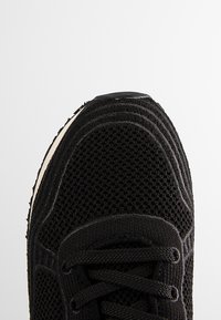 Czarne oddychające sneakersy z siateczki, z wypukłym przeszyciem i czarnymi sznurowadłami, widziane z góry, na jednolitym białym tle.