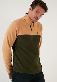 Flis jakna s polu-zip dizajnom, svijetlosmeđeg gornjeg dijela i tamnozelenog donjeg dijela. Mekana tekstura i mali logo na prsima.