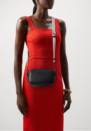 Kobieta nosząca czerwoną sukienkę bez rękawów z kwadratowym dekoltem, niosąca czarną torebkę na ramię z beżowym paskiem i nosząca złotą bransoletkę łańcuchową.