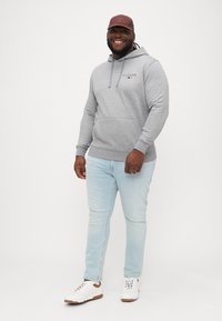 Šedá mikina s nastaviteľnými šnúrkami a predným vreckom, svetlomodré džínsy a biele tenisky s hnedými akcentmi. Čiapka v tmavočervenej farbe.