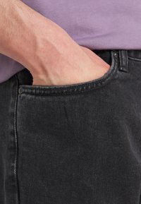 Odrasla roka vstavljena v sprednji žep črnih kavbojk, s poudarjeno purpurno srajco, delno vidno nad pasom.