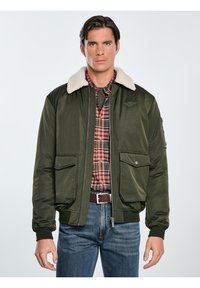 Grüne Bomberjacke mit beigem Kunstfellkragen, Reißverschluss vorne und zwei großen vorderen Taschen. Getragen über einem karierten Hemd und Jeans.