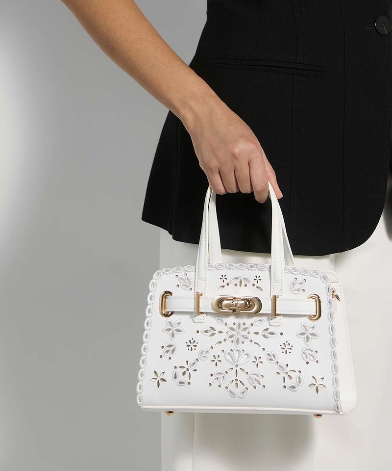 Main tenant un sac à main blanc avec des motifs floraux découpés et un fermoir doré, portant un haut noir et un pantalon blanc sur un fond gris.
