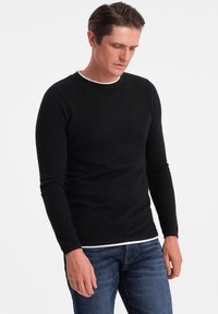 Black long-sleeve sweater with a textured pattern, round neckline, and white trim. Paired with blue jeans, the fit is slim and casual.