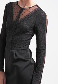 Top de encaje negro con mangas largas transparentes, escote en V, patrón floral y una falda de satén ajustada. Diseño elegante con tela texturizada.