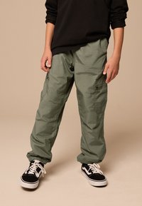 Pantalon cargo vert olive, avec des poignets fuselés, plusieurs poches, et un matériau léger et texturé associé à des baskets noires.