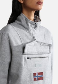 Pullover in pile grigio chiaro con colletto alto, tasca frontale con zip e una toppa con la bandiera norvegese. Texture morbida e design sportivo.
