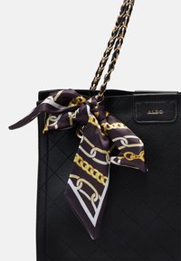 Bolso tote de cuero sintético acolchado negro con correa de cadena en tono dorado y una bufanda de seda atada al asa, que presenta un patrón de cadenas en negro y dorado.