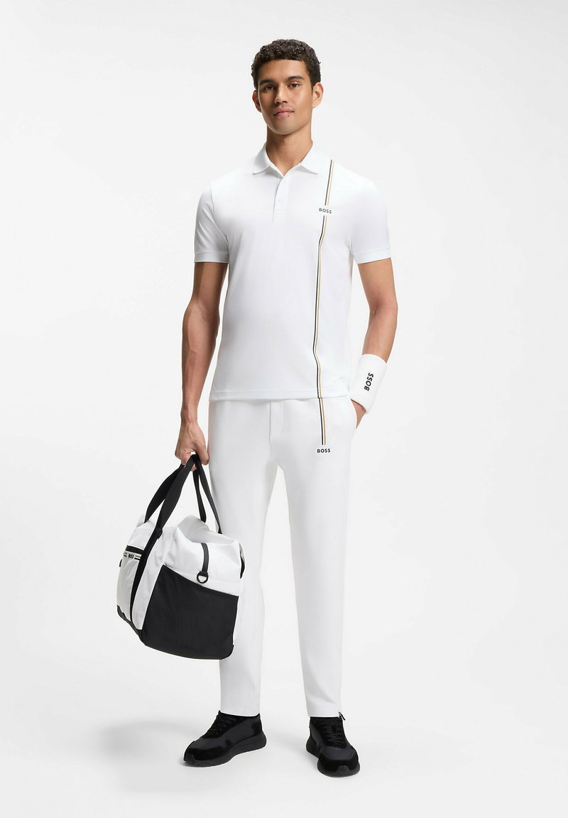 Homme portant un polo blanc et un pantalon avec des rayures noires et dorées, tenant un sac polochon blanc et noir, debout sur un fond blanc.