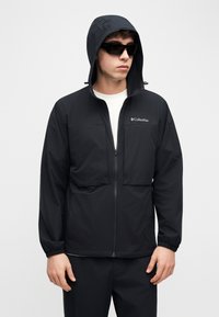 Άνδρας που φοράει μαύρο μπουφάν Columbia με κουκούλα, λευκό πουκάμισο, μαύρο παντελόνι και μαύρα γυαλιά ηλίου μπροστά σε απλό φωτεινό φόντο.