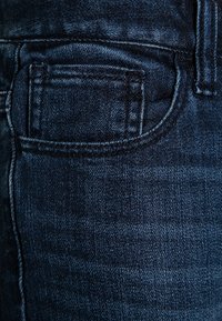 Tmavě modré džíny vyrobené z denimu, které mají zadní kapsu s viditelným prošíváním a hladkou texturou. Tkanina má lehký lesk.