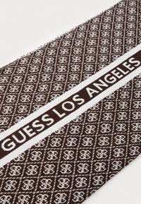 Brun sjal med et hvitt mønster og bold tekst "GUESS LOS ANGELES." Stoffet har en teksturert overflate med et diamantliknende design gjennomgående.