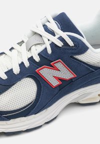 Gros plan d'une basket bleu marine et blanche avec des lacets blancs et un logo gris "N" entouré de rouge sur le côté.