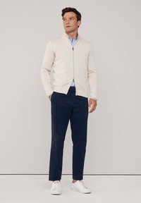 Suéter de cremallera color beige claro con un dobladillo acanalado, combinado con pantalones oscuros de color marino y zapatillas blancas. Lleva una camisa de rayas azules debajo.