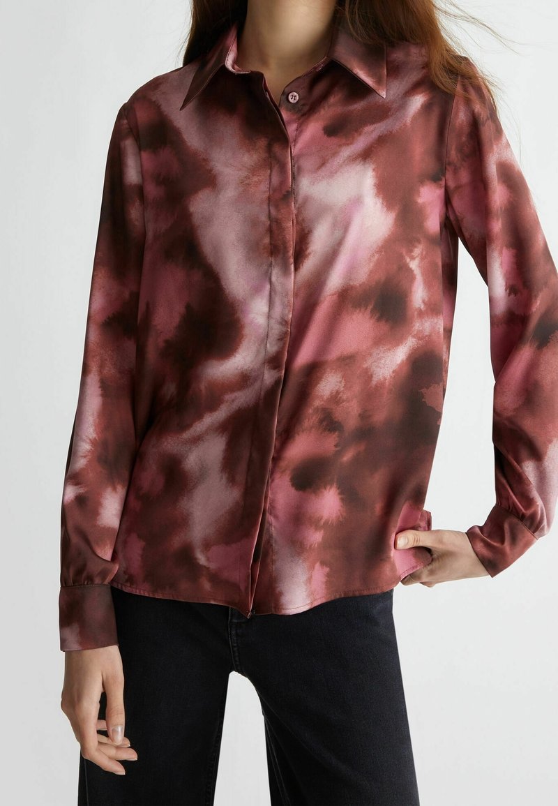 Femme portant une chemise à manches longues boutonnée avec un motif tie-dye rouge et noir, associée à un pantalon noir, main sur la hanche.