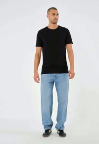 T-shirt nera a maniche corte abbinata a jeans azzurri, indossata con scarpe da ginnastica nere, che presenta un outfit casual su uno sfondo neutro.