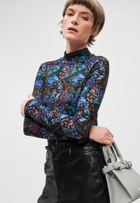 Langärmliges Oberteil mit einem Blumenmuster in Blau, Grün und Lila auf schwarzem Hintergrund; kombiniert mit hochgeschnittenen schwarzen Hosen und einer grauen Tasche.