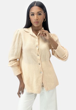 Donna con lunghi capelli scuri, orecchini a cerchio dorati e camicia beige testurizzata con bottoni e maniche a sbuffo, abbinata a pantaloni bianchi.