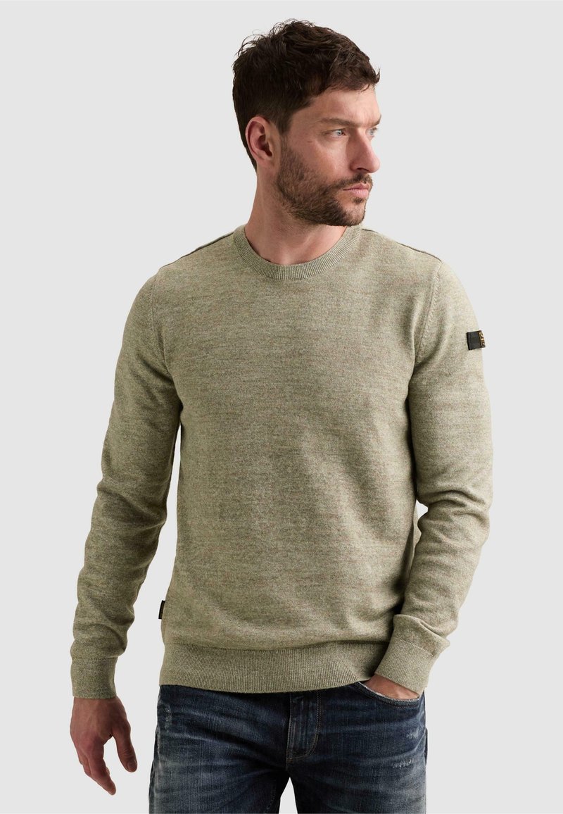 Homme portant un pull col rond vert olive clair et un jean bleu, regardant vers sa droite avec une main dans la poche sur fond uni.