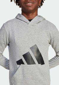 Felpa grigia con cappuccio e tasca frontale, caratterizzata da un grande logo Adidas nero posizionato in basso a destra. Realizzata in un morbido tessuto testurizzato.