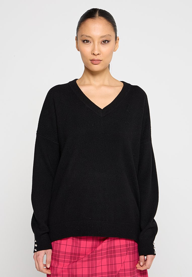 Maglione nero oversize con scollo a V, texture a coste e maniche lunghe con dettagli a bottone sui polsini. Indossato sopra una gonna a quadri rosa.