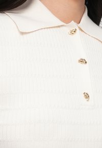 Biela prúžkovaná polokošeľa s prekladaným golierom a tromi zlatými gombíkmi s dekoratívnym vzorom. Textúra je hladká a priliehavá.