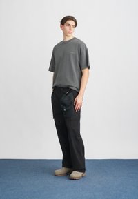 Γκρίζα oversized t-shirt, μαύρο cargo παντελόνι με τσέπες, μπεζ slip-on παπούτσια και ένα μαύρο καπέλο που κρατιέται στο χέρι, σε απλό φόντο.