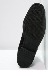 Semelle de chaussure en daim noir présentant un dessous en caoutchouc texturé avec un motif géométrique et des détails de marque, inclinée sur un fond blanc.