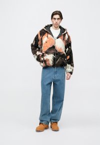 Giacca morbida e multicolore con motivi arancioni, neri e crema, dotata di cerniera frontale e cappuccio, abbinata a jeans larghi blu e scarpe color sabbia.