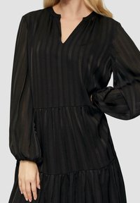 Černé dlouhé šaty s dlouhým rukávem a svislými pruhy, výstřihem do „V“, nařaseným živůtkem a stupňovitou sukní; vyrobené z lehké texturované látky.