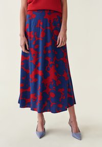Vrouw in een blauw en rood bloemig midi-jurk, rode top en blauwe pointy-toe hakken, staand tegen een effen lichte achtergrond.