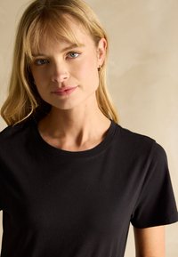 Blonde woman with blue eyes wears small gold hoop earrings and a black crew neck t-shirt, posing against a plain beige background.