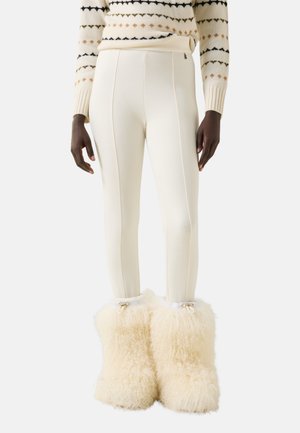 Persoon draagt crèmegekleurde aangesloten broek, een patroontrui en pluizige crèmegekleurde winterlaarzen op een witte achtergrond.